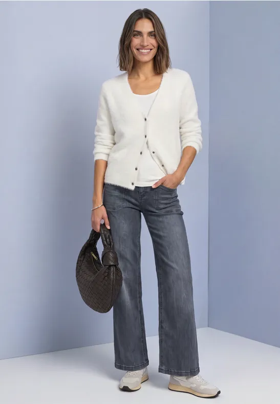 Frau mit weißer Strickjacke, grauen Jeans, Sneakern und runder Korbtasche.