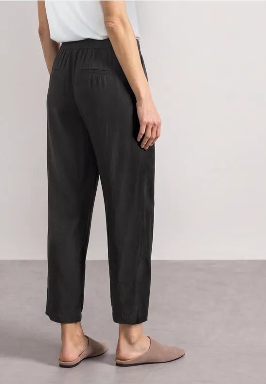 Ein Modell von hinten in schwarzen, lässigen, hoch geschnittenen Hosen mit elastischem Bund und beigen Slippern.