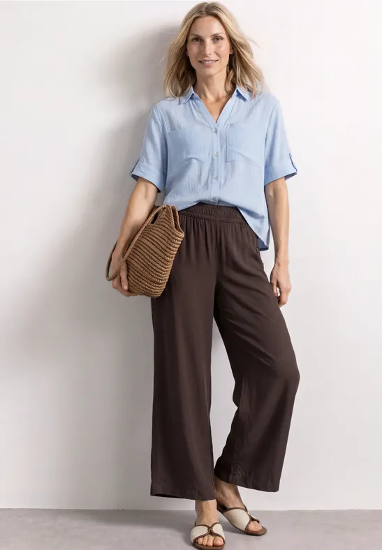 Frau in hellblauer Bluse und weiten braunen Hosen mit Basttasche und Sandalen vor neutralem Hintergrund.