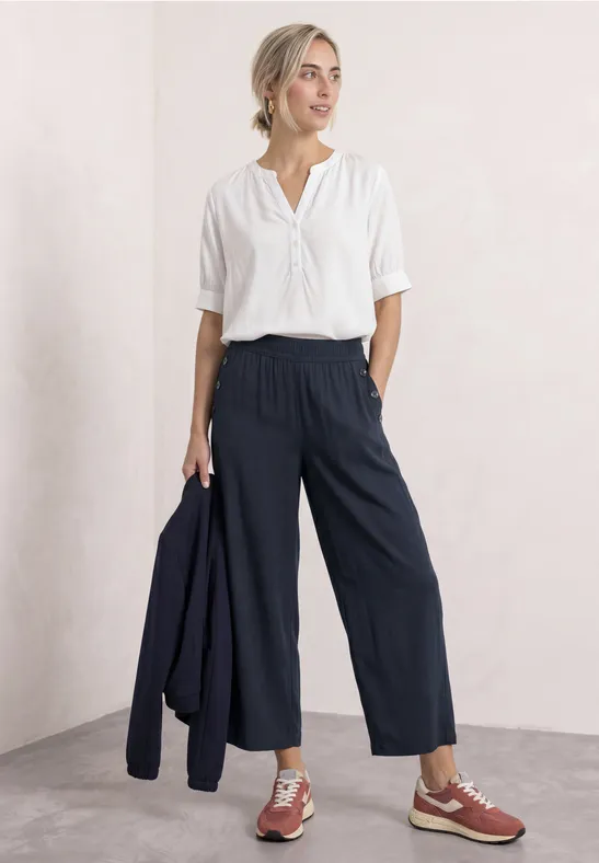 Frau in weißer Bluse und dunkelblauen Culottes hält eine Jacke, trägt braune Schuhe.