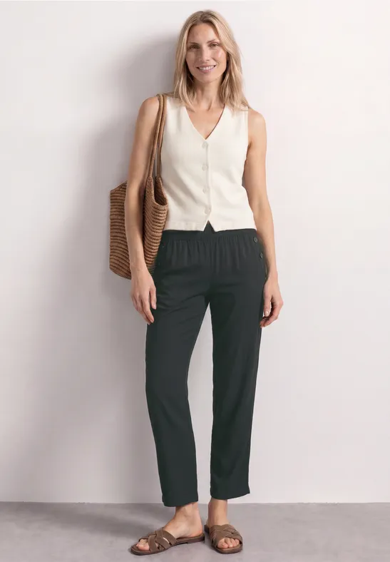 Eine Frau trägt ein weißes ärmelloses Top, schwarze Hose und Sandalen und hält eine geflochtene Tasche.