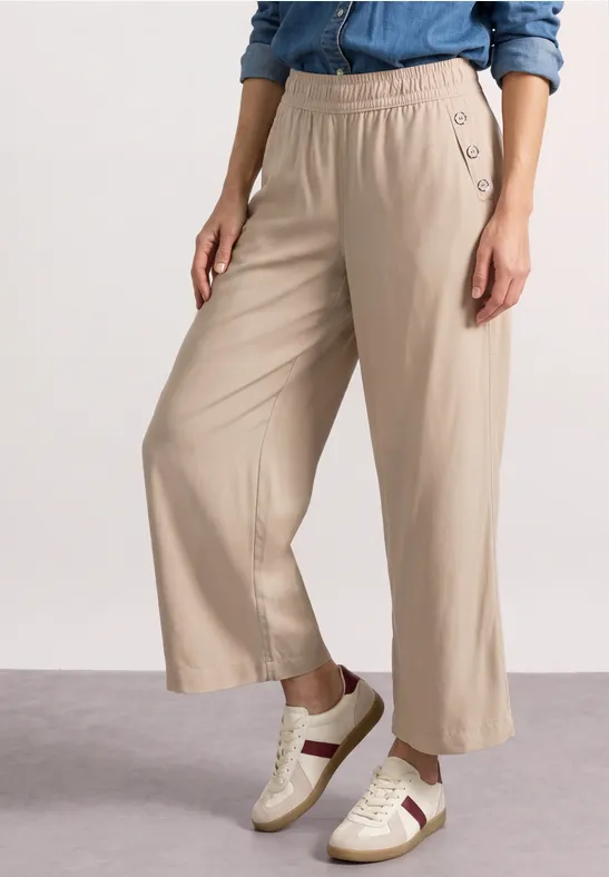 Dame draagt een beige wijdvallende broek met elastische taille en knopen, gecombineerd met witte sneakers met strepen.