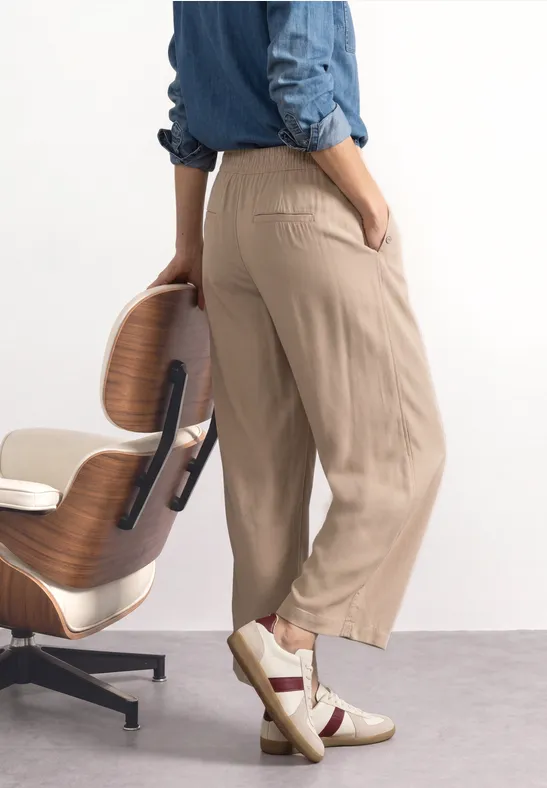 Een persoon draagt beige broek en witte sneakers en leunt tegen een houten stoel.
