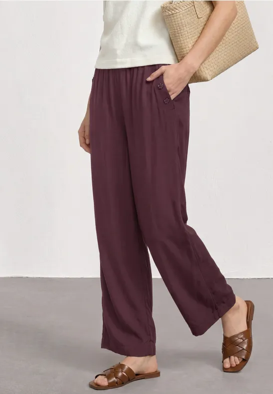 Eine Person trägt weiche, locker sitzende bordeauxrote Hose mit Taschen, braune Sandalen und Korbtasche.