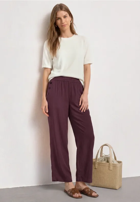 Dame trägt weißes T-Shirt und weinrote Hose, Sandalen, neben ihr eine Basttasche.