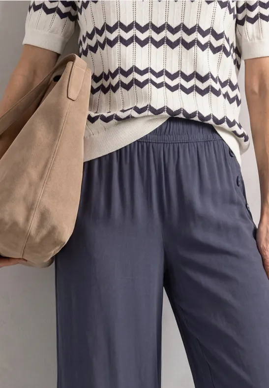 Eine Person trägt ein weiß-blaues, zickzackgemustertes Strickshirt, dunkelblaue Freizeithose und eine hellbraune Tasche.