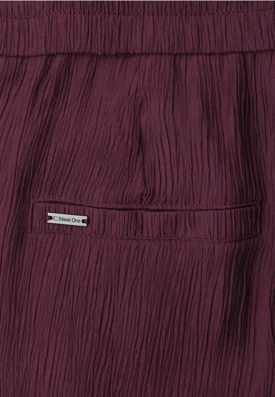 Nahaufnahme einer weinroten, plissierten Hose mit Gesäßtasche und kleinem Metall-Emblem