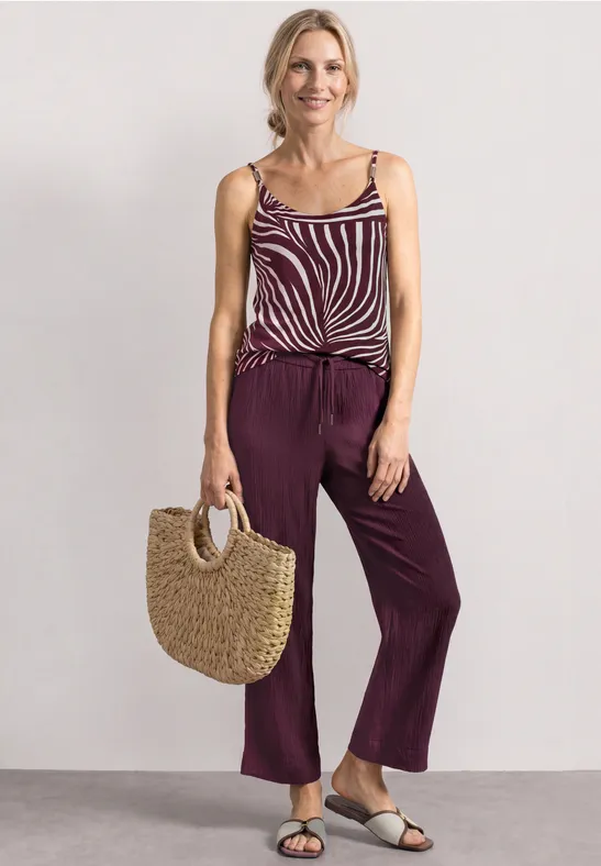 Frau trägt gestreiftes Top, breite lila Hose, Sandalen und eine Strohtasche.