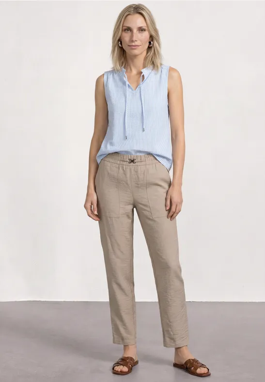 Frau trägt hellblaues ärmelloses Top, beige Hose und braune Sandalen vor schlichtem Hintergrund.