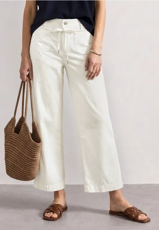 Frau in weißen, weiten Culottes mit braunen Sandalen, schwarzem Top und geflochtener Strohtasche.