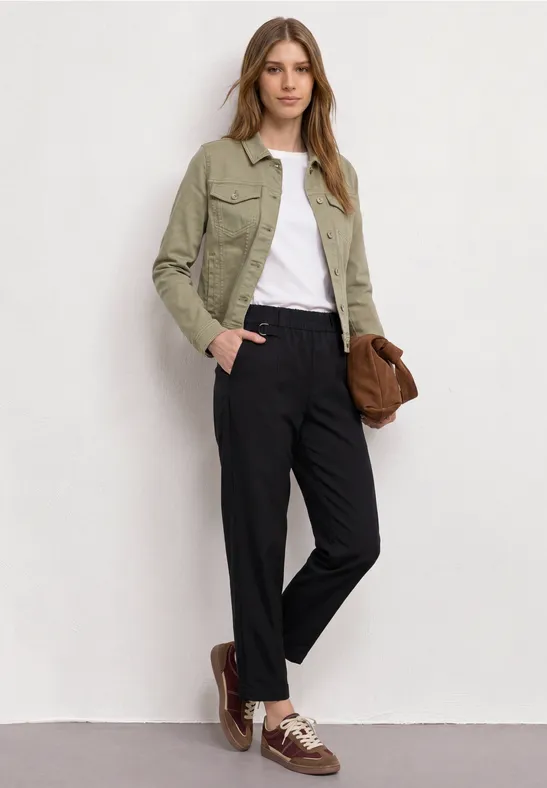 Persoon voor witte muur met groene jas, wit shirt, zwarte broek, bruine schoenen en tas.