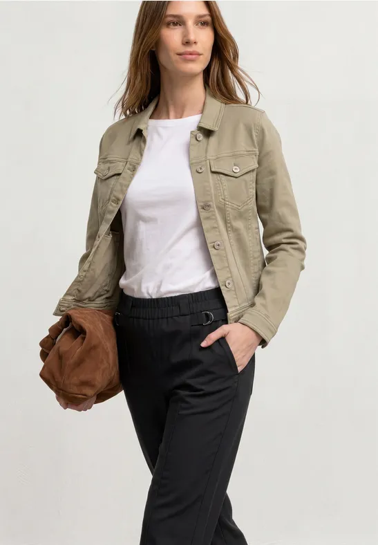 Vrouw in lichtbeige jack, wit T-shirt en zwarte broek, houdt een bruine tas, hand in de zak.