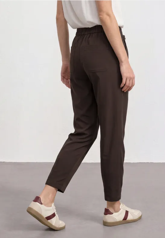 Vrouw van achteren in een losvallende bruine broek, wit hemdje en witte sneakers voor een neutrale achtergrond.