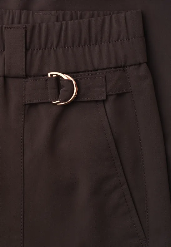 Close-up van een donkerbruine broek met elastische taille, steekzak en goudkleurige D-ring gesp.