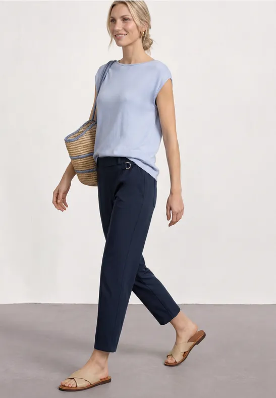 Vrouw in lichtblauw topje, donkerblauwe broek, platte sandalen en een rieten tas.
