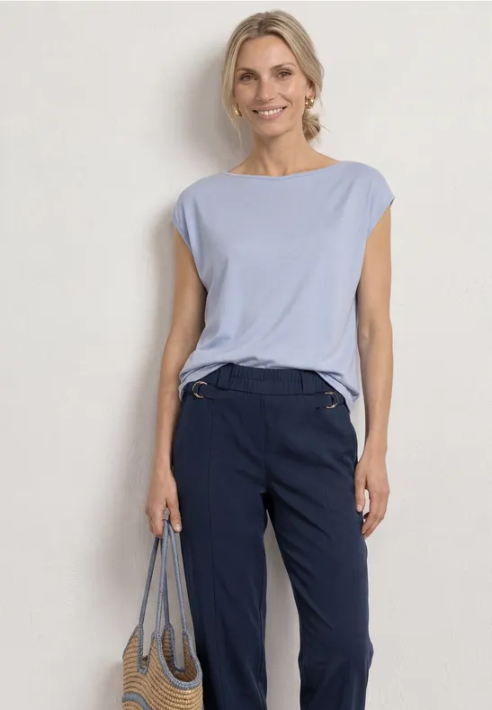 Vrouw voor witte muur met lichtblauw shirt, donkerblauwe broek en gestreepte tas.