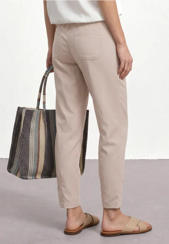 Achteraanzicht van persoon in beige broek en slippers, met een gestreepte tas in de hand.