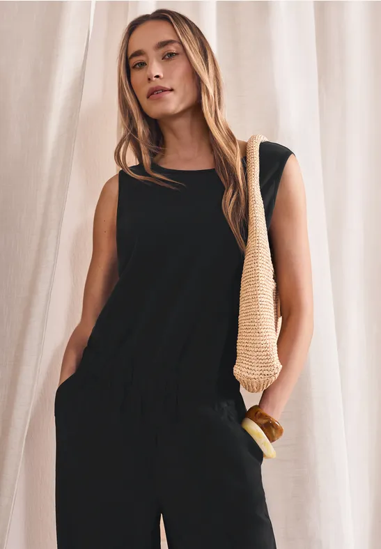 Vrouw draagt zwarte mouwloze jumpsuit met elastische taille en zakken, plus een gevlochten schoudertas.