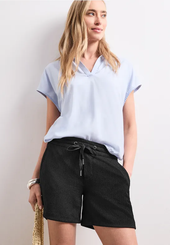 Vrouw in lichtblauw bloesje, zwarte short en met strooien tas.