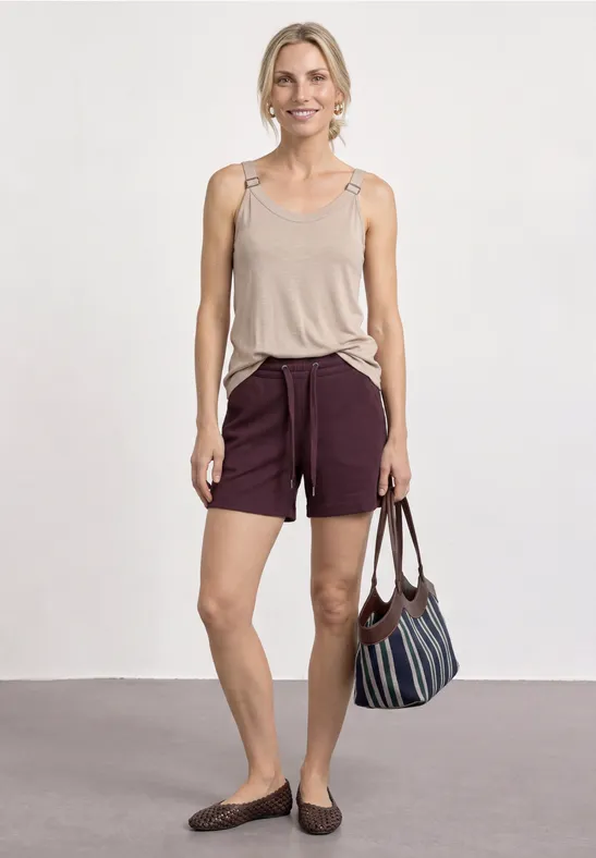 Vrouw voor witte muur, draagt beige top, bordeaux short, ballerina’s en houdt een gestreepte tas vast.