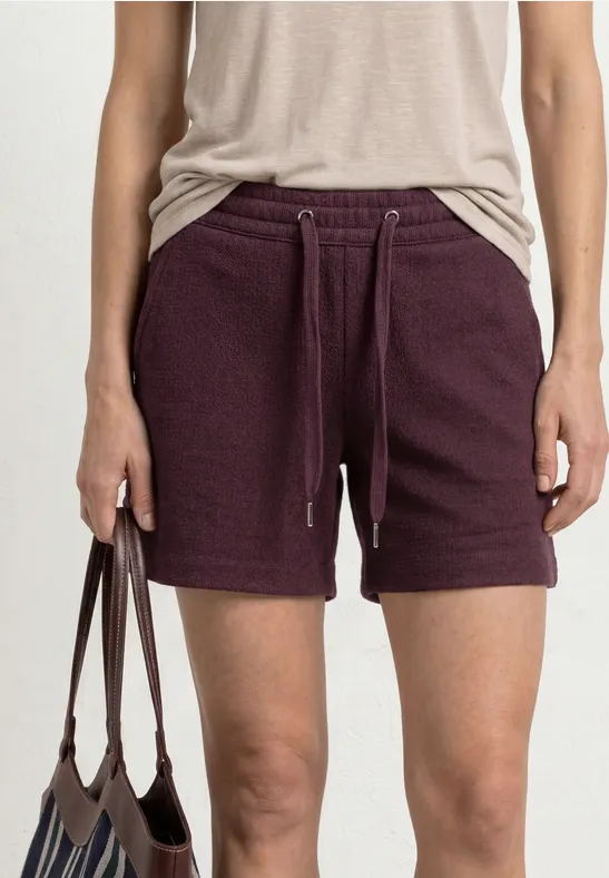 Persoon draagt een beige top en donkerpaarse sweatshorts met trekkoord, houdt een bruine tas vast.