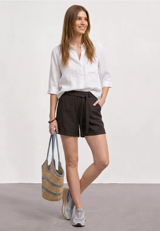 Eine Frau trägt eine weiße Bluse, schwarze Shorts, graue Sneaker und hält eine gestreifte Strohtasche.
