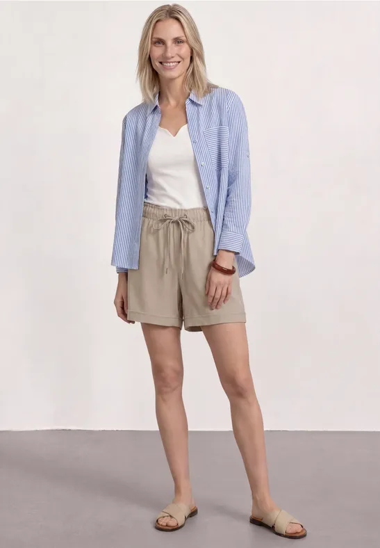 Frau trägt beige Shorts, weißes T-Shirt, hellblaues Hemd und Sandalen vor hellem Hintergrund.