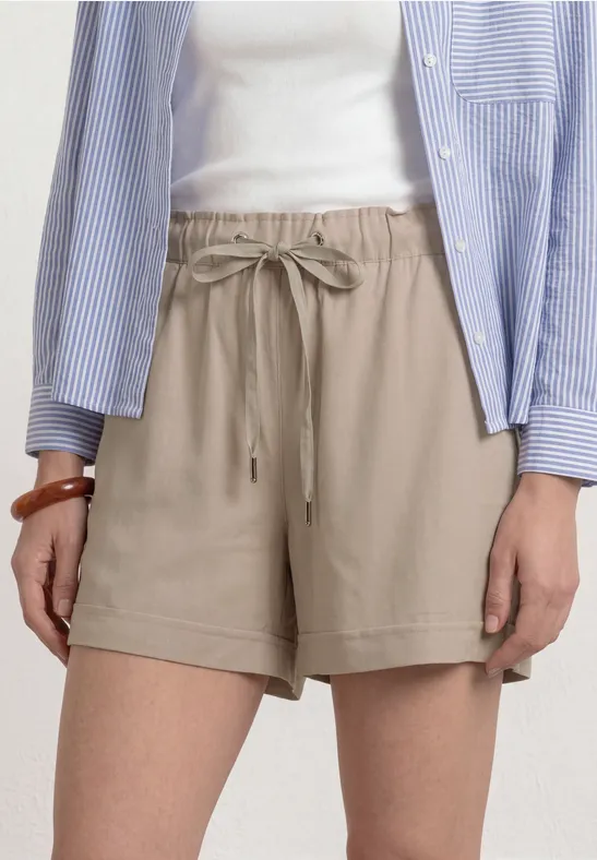 Beige Damen-Shorts mit Tunnelzug, kombiniert mit weißem Top und blau-weiß gestreifter Hemdbluse.