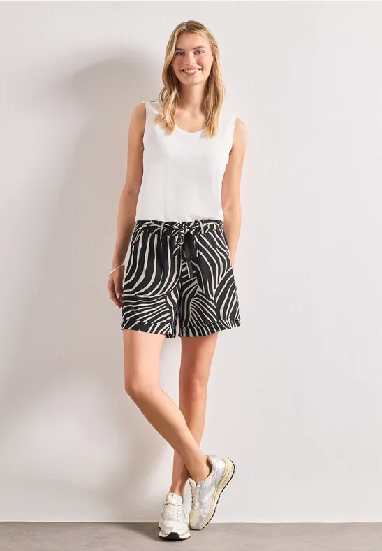 Une jeune femme pose devant un mur blanc avec un débardeur blanc, un short zébré et des baskets.