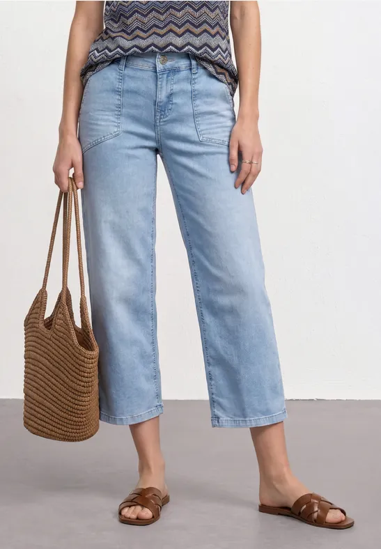 Person trägt hellblaue, gerade geschnittene Jeans, gemustertes Top, Basttasche und geflochtene Sandalen.