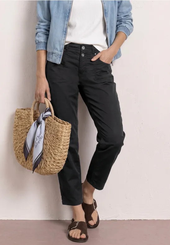 Frau trägt schwarze Hose, weißes Top, Jeansjacke, Basttasche mit Tuch und braune Sandalen.