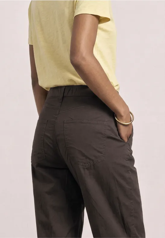 Rückenansicht einer Person in gelbem Oberteil und dunkelbrauner Hose, Hände in den Taschen.