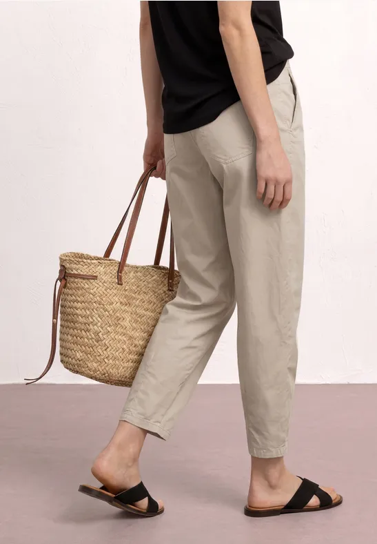 Person trägt beige Hose, schwarze Sandalen und hält einen geflochtenen Strohkörben.