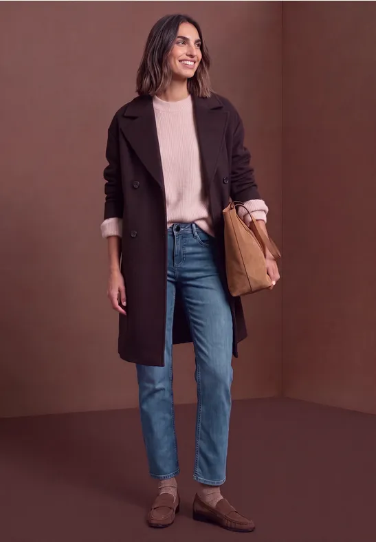 Une femme souriante en manteau marron, pull beige, jean et chaussures brunes devant un fond brun.