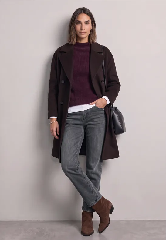 Frau im dunkelbraunen Mantel, bordeauxrotem Pullover über weißer Bluse, grauen Jeans, braunen Stiefeletten und schwarzer Tasche.