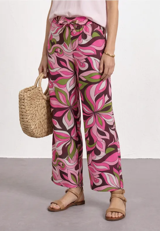 Vrouw draagt ruime roze broek met abstract bladprint, afgewerkt met strozak en sandalen.