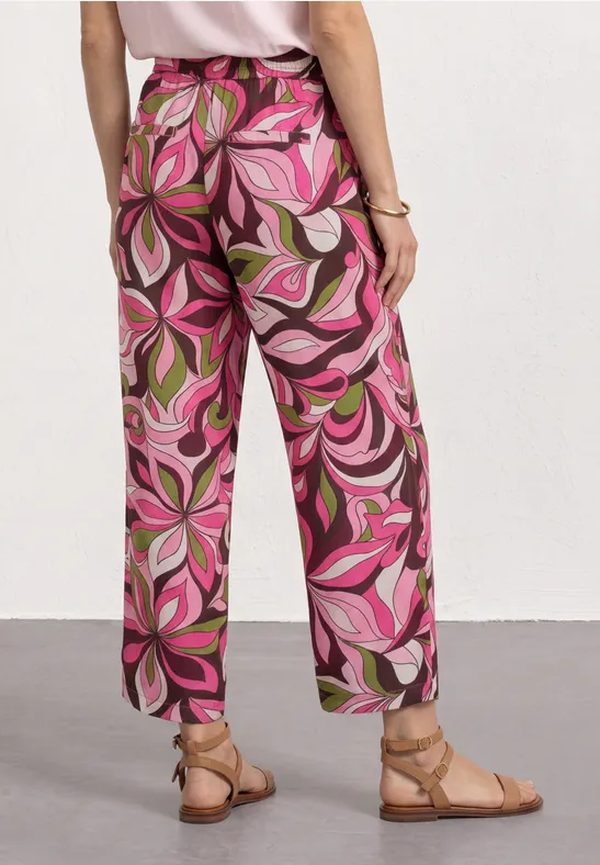 Persoon draagt een broek met sierlijke roze-groene print en platte sandalen voor een witte achtergrond.