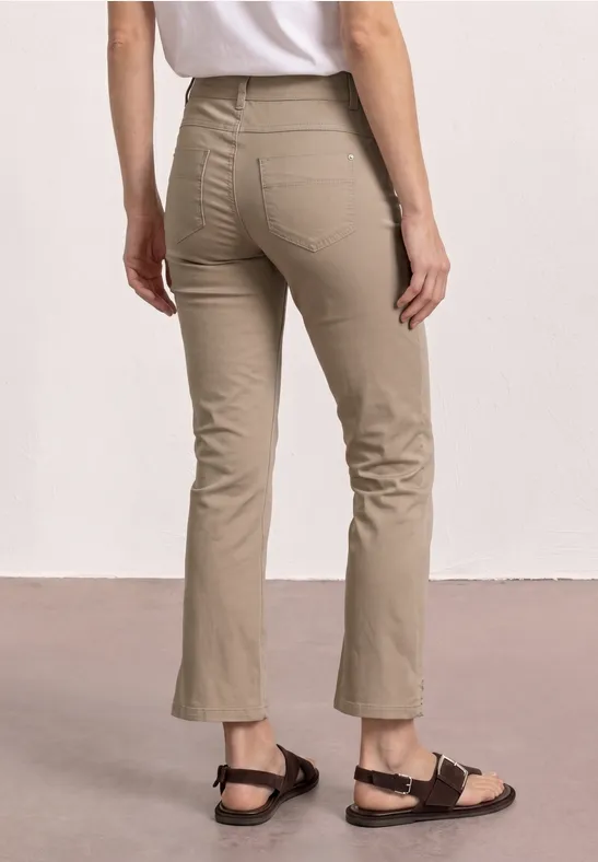 Vrouw van achter in licht beige, licht uitlopende 7/8-broek en zwarte sandalen tegen neutrale achtergrond.