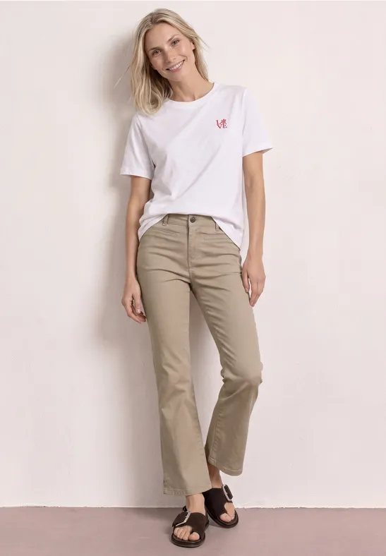 Vrouw leunt nonchalant tegen lichte muur, draagt een wit t-shirt, beige wijde 7/8-broek en zwarte sandalen.