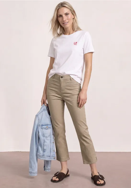 Een vrouw draagt een wit T-shirt met een rood hartje, beige broek, zwarte sandalen en houdt een spijkerjack vast.