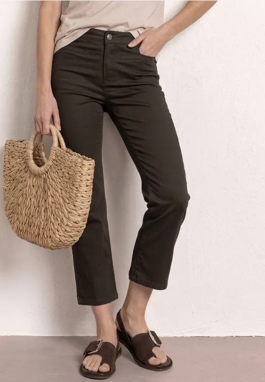 Persoon draagt zwarte cropped broek, houdt een gevlochten rieten tas en draagt bruine sandalen.