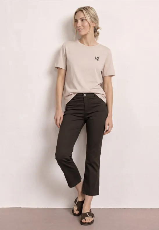 Vrouw in beige T-shirt, zwarte 7/8-broek en sandalen voor een neutrale achtergrond.