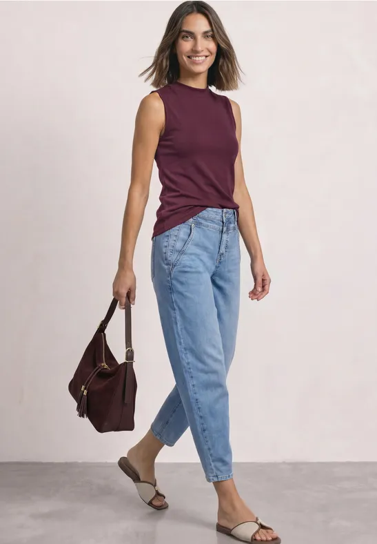 Frau in Jeans, ärmellosem dunkelrotem Top, Sandalen und Tasche, die lächelnd posiert.