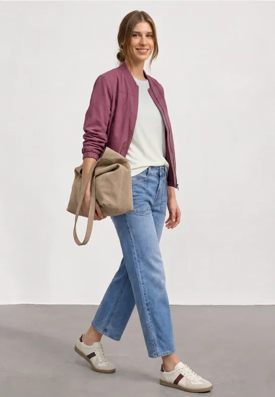 Frau in lässigem Outfit: bordeauxroter Blouson, weißes Shirt, gerade geschnittene Jeans, weiße Sneaker und Beuteltasche.