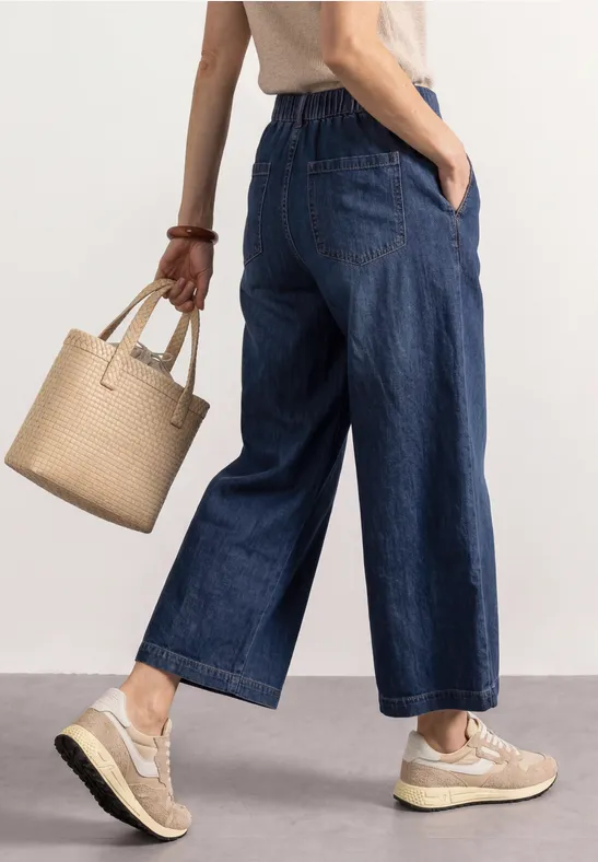 Vrouw in wijde, korte spijkerbroek, beige sneakers en rieten tas tegen lichte achtergrond.