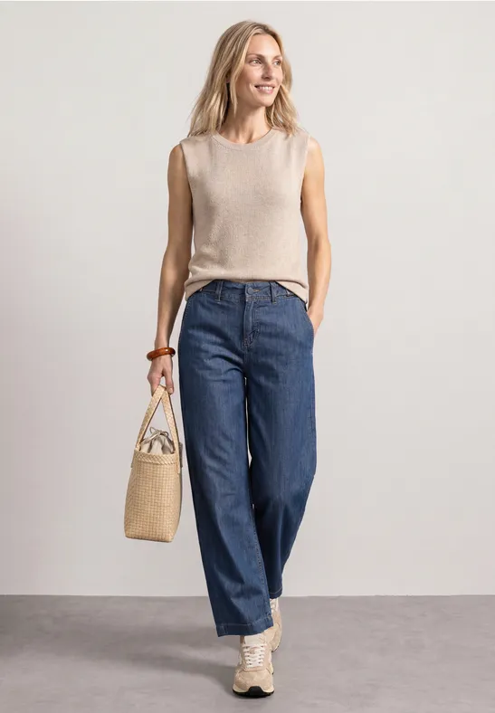 Een vrouw draagt een beige top, wijde blauwe jeans en sneakers, met een strooien tas voor een lichte achtergrond.