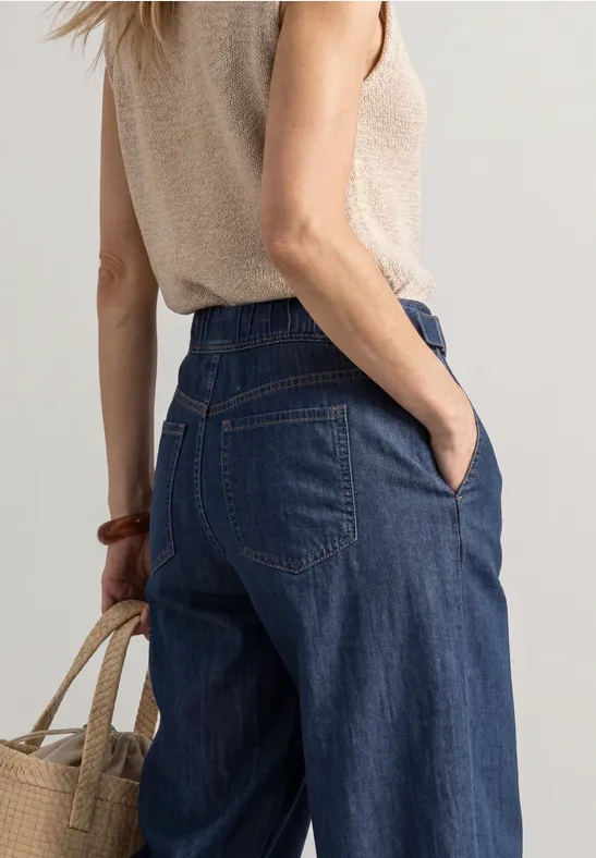 Vrouw van achteren in wijde jeans en beige top, ze draagt een strooien tas.
