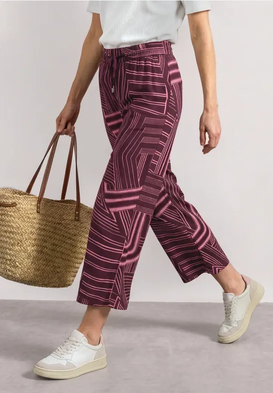 Femme en t-shirt blanc, pantalon rose à motifs, baskets blanches et sac en osier.