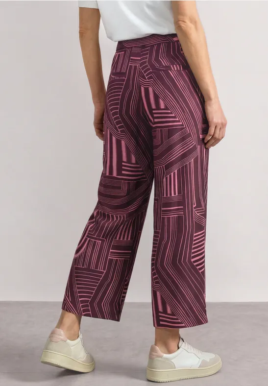 Une femme porte un pantalon bordeaux ample à motifs géométriques, associé à un t-shirt blanc et des baskets.