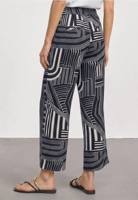 Wijde broek met zwart-beige geometrisch strepenpatroon en gouden sandaaltjes met bandjes.